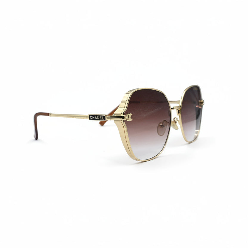 Oversized square sunglasses with a gold metal frame and brown gradient lenses, displayed on a white surface with a reflective silver backdrop.