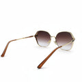 Oversized square sunglasses with a gold metal frame and brown gradient lenses, displayed on a white surface between two silver reflective fabric panels.