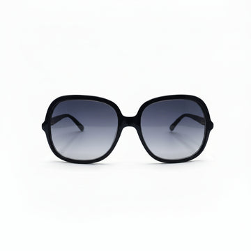 Oversized square sunglasses with a thick black frame and dark gradient lenses, displayed on a white surface with two textured silver props on either side.
