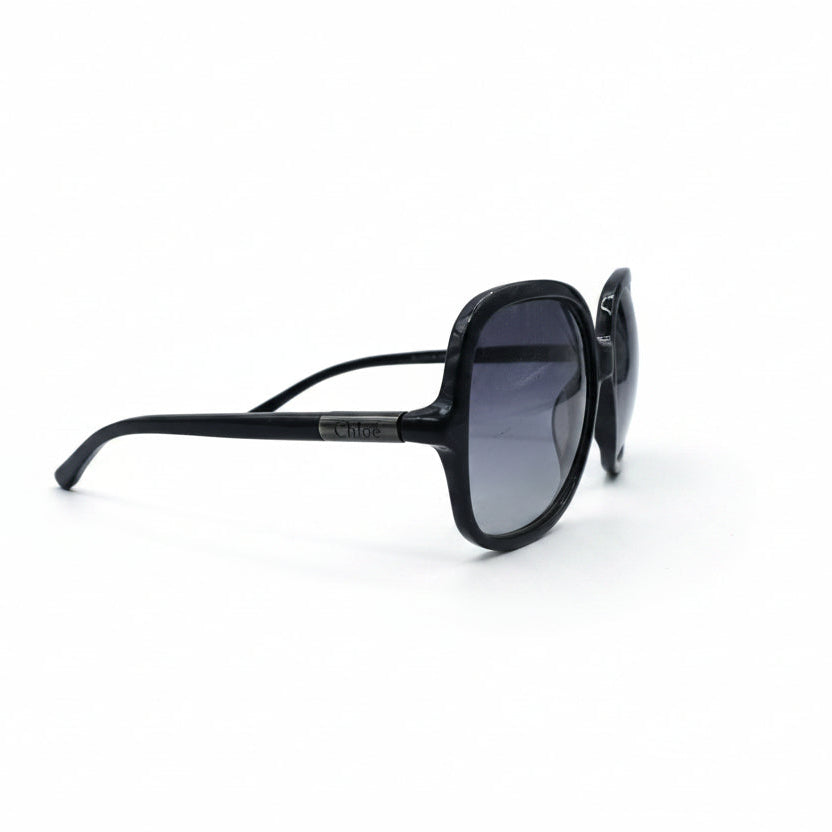 Oversized square sunglasses with a thick black frame and dark gradient lenses, displayed on a white surface with two textured silver props on either side.