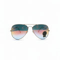 A pair of aviator sunglasses with a gold metal frame and gradient lenses shifting from teal-blue to pink-orange, displayed on a white surface between two textured silver panels.