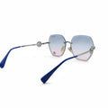 Oversized square sunglasses with a silver-tone metal frame, gradient blue-to-pink lenses, and blue temple tips, displayed on a white surface between two silver fabric panels.