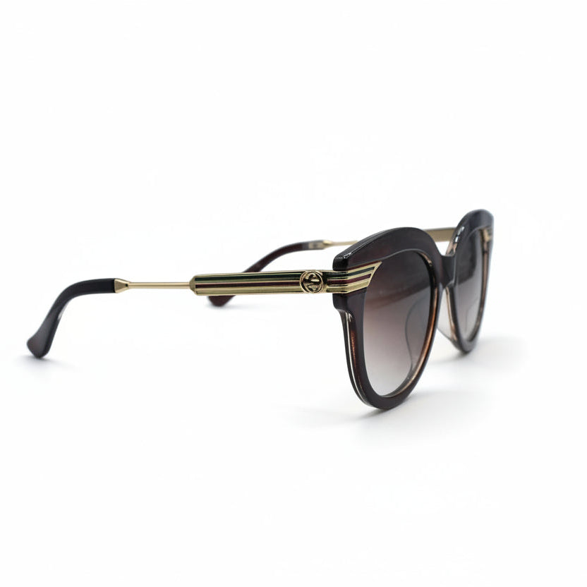Gucci cat-eye sunglasses with a translucent dark brown frame and gradient brown lenses, featuring gold metal double stripes on the temples and the Gucci logo on the lens.