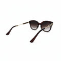 Oversized cat-eye sunglasses with dark brown gradient lenses and gold metal temples, displayed on a white surface with textured silver panels in the background.