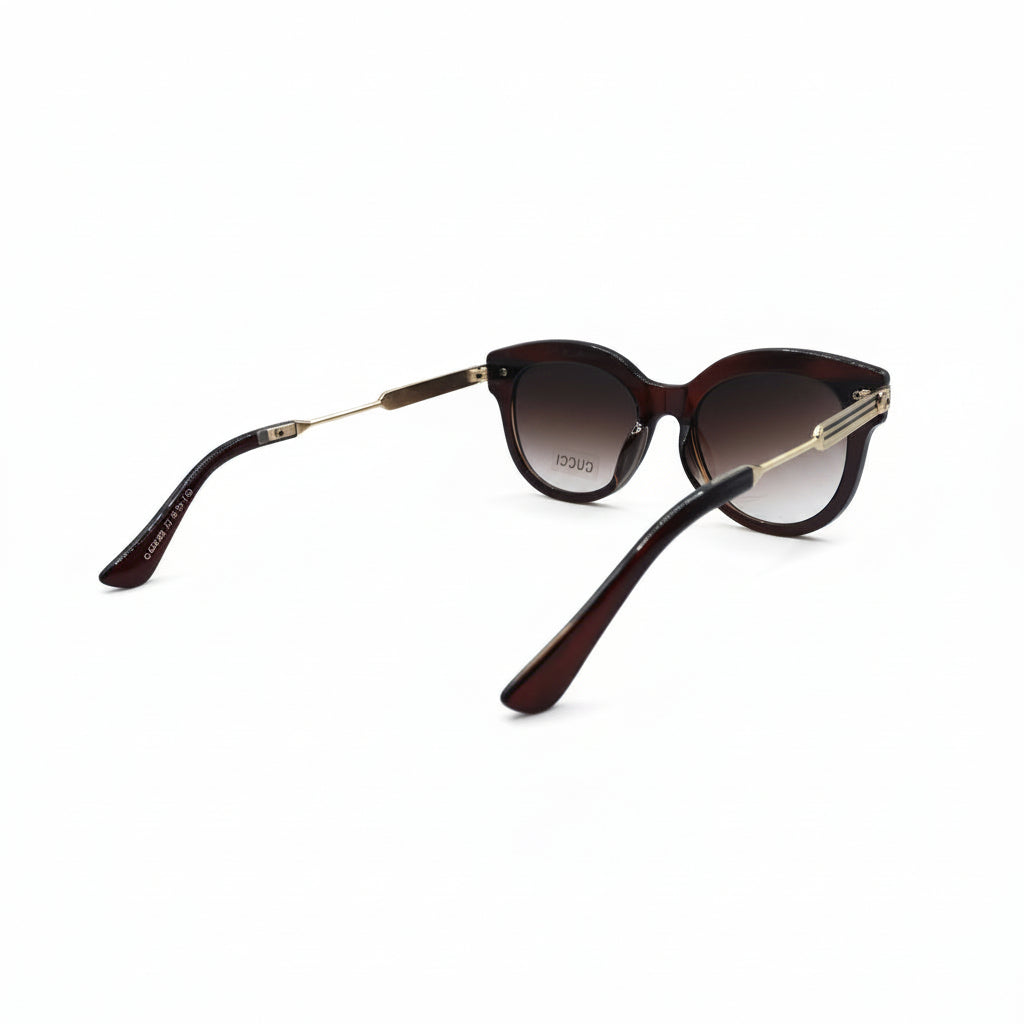 Oversized cat-eye sunglasses with dark brown gradient lenses and gold metal temples, displayed on a white surface with textured silver panels in the background.