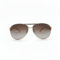 A pair of classic aviator sunglasses with a gold metal frame and brown gradient lenses, displayed on a white surface with a gray textured backdrop on either side.