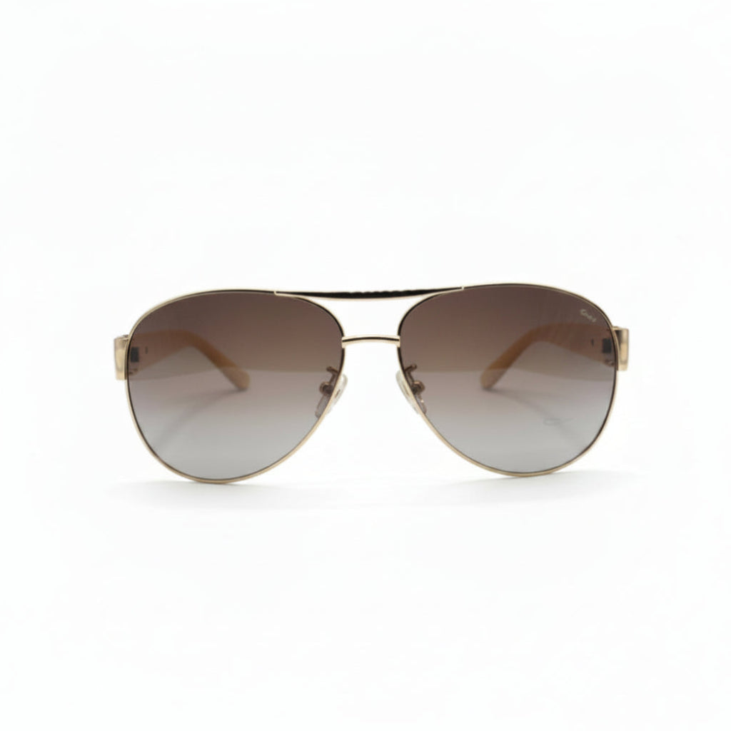 A pair of classic aviator sunglasses with a gold metal frame and brown gradient lenses, displayed on a white surface with a gray textured backdrop on either side.