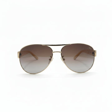A pair of classic aviator sunglasses with a gold metal frame and brown gradient lenses, displayed on a white surface with a gray textured backdrop on either side.