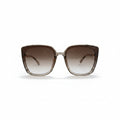 A pair of oversized square sunglasses with a translucent taupe-brown acetate frame and brown gradient lenses, displayed on a white surface with two textured silver props on either side.