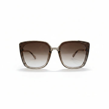 A pair of oversized square sunglasses with a translucent taupe-brown acetate frame and brown gradient lenses, displayed on a white surface with two textured silver props on either side.