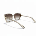 Oversized square sunglasses with a translucent champagne-gold acetate frame and brown gradient lenses, displayed on a white surface between two textured gray panels.