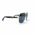 A pair of aviator-style sunglasses with a gold metal frame, black temples, and dark blue-gray tinted lenses, displayed on a white surface with reflective silver studio lights in the background.