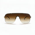 A pair of oversized shield-style sunglasses with a gradient brown lens and gold metal frames, displayed on a white studio background with textured silver props on either side.