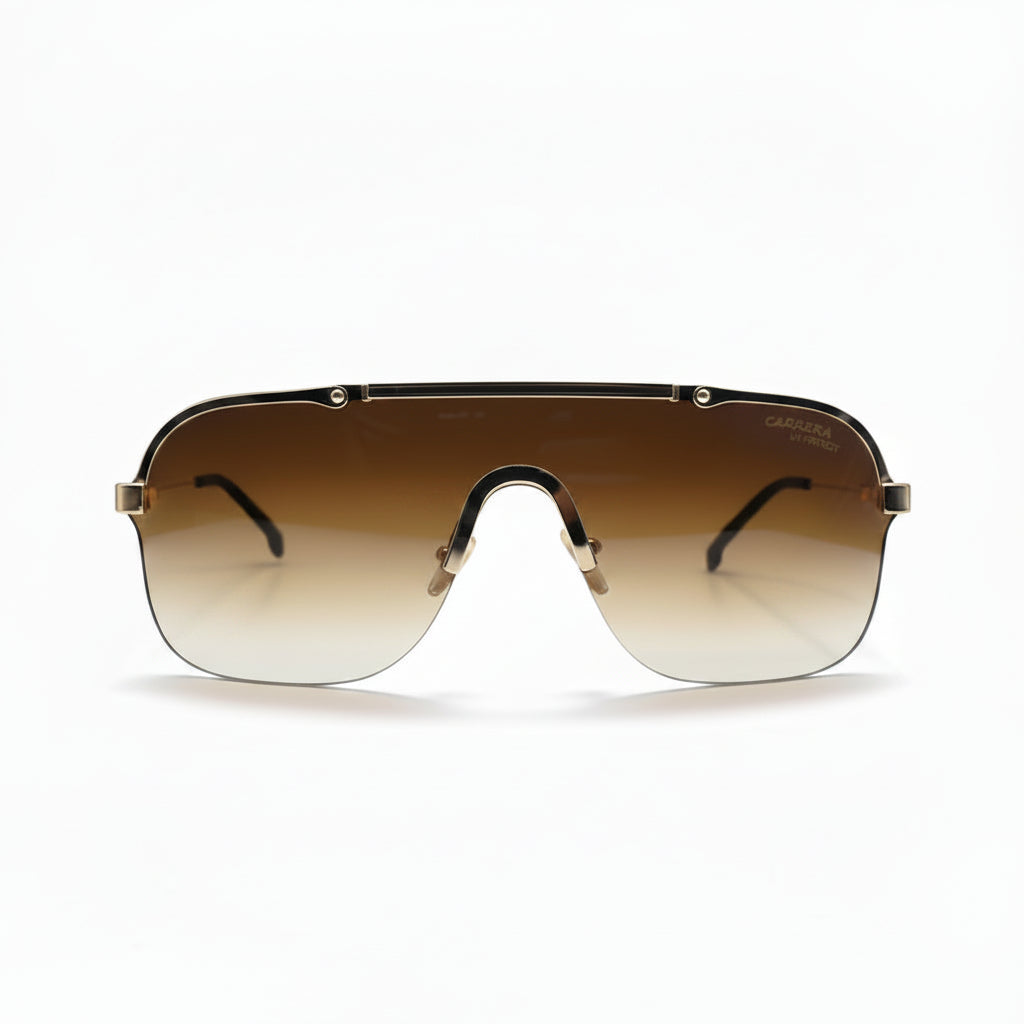 A pair of oversized shield-style sunglasses with a gradient brown lens and gold metal frames, displayed on a white studio background with textured silver props on either side.