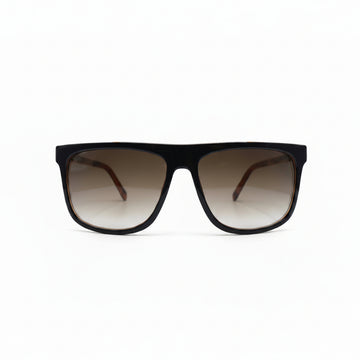 A pair of square-frame sunglasses with a thick black frame and brown gradient lenses, displayed on a white surface with two black fabric panels in the background.
