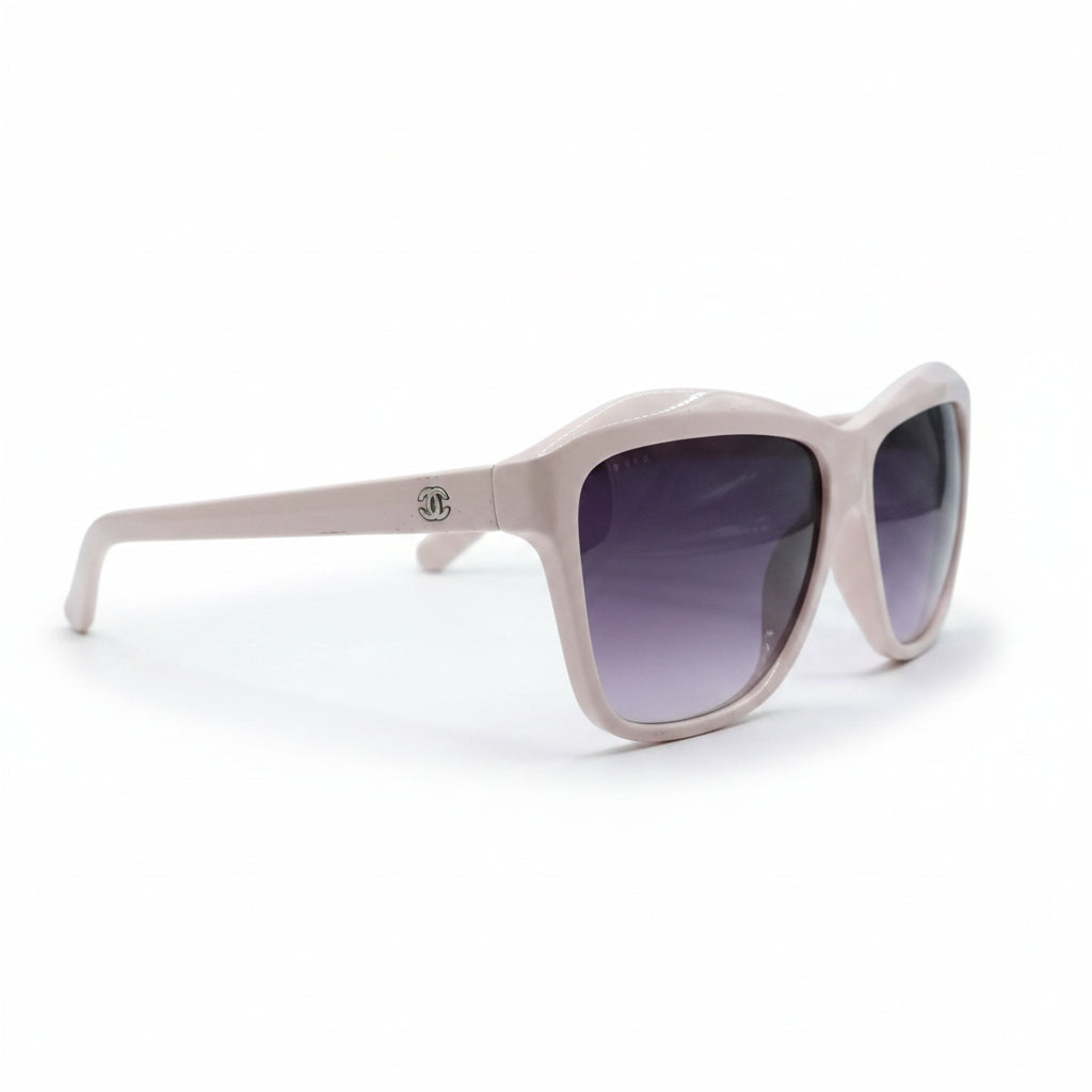 A pair of oversized square sunglasses with a light pink frame and dark purple gradient lenses, displayed on a white surface with soft studio lighting.