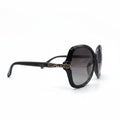 A pair of designer sunglasses with a dark brown acetate frame, gold-tone ornate details on the temples, and dark gray gradient lenses, displayed on a white surface with studio lights in the background.