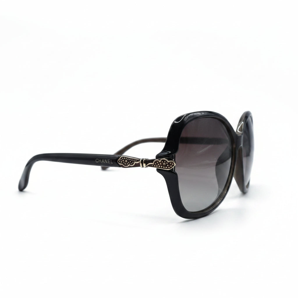 A pair of designer sunglasses with a dark brown acetate frame, gold-tone ornate details on the temples, and dark gray gradient lenses, displayed on a white surface with studio lights in the background.