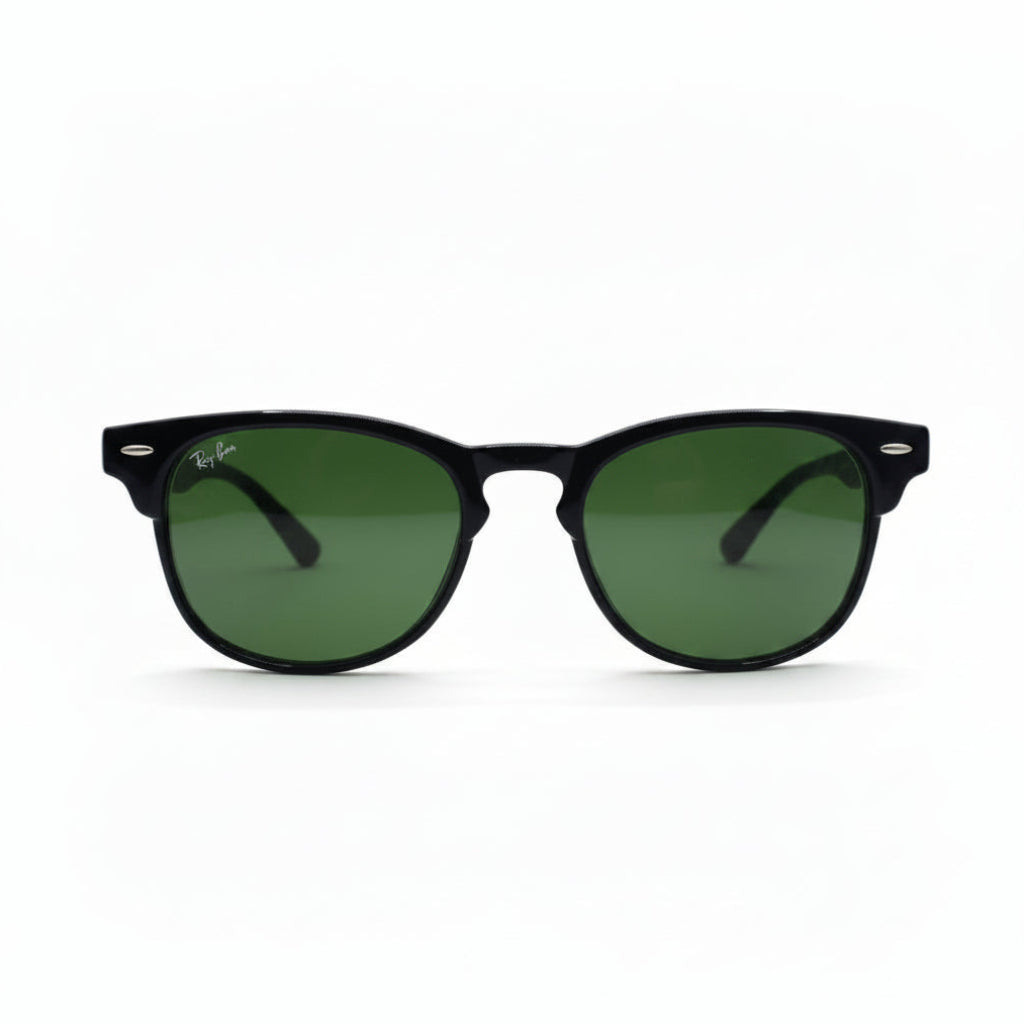 Black classic-style sunglasses with green lenses, featuring a visible Ray-Ban logo sticker on the lens, set against a white background with textured gray props on either side.