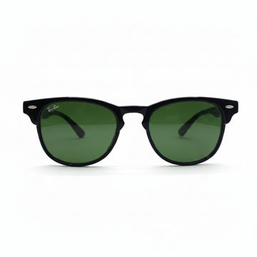 Black classic-style sunglasses with green lenses, featuring a visible Ray-Ban logo sticker on the lens, set against a white background with textured gray props on either side.