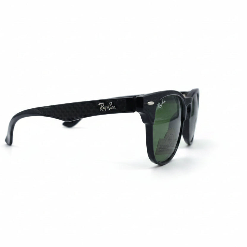 Black classic-style sunglasses with green lenses, featuring a visible Ray-Ban logo sticker on the lens, set against a white background with textured gray props on either side.