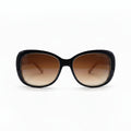 A pair of oversized cat-eye sunglasses with a glossy black frame and brown gradient lenses, displayed on a white surface with a reflective silver backdrop.