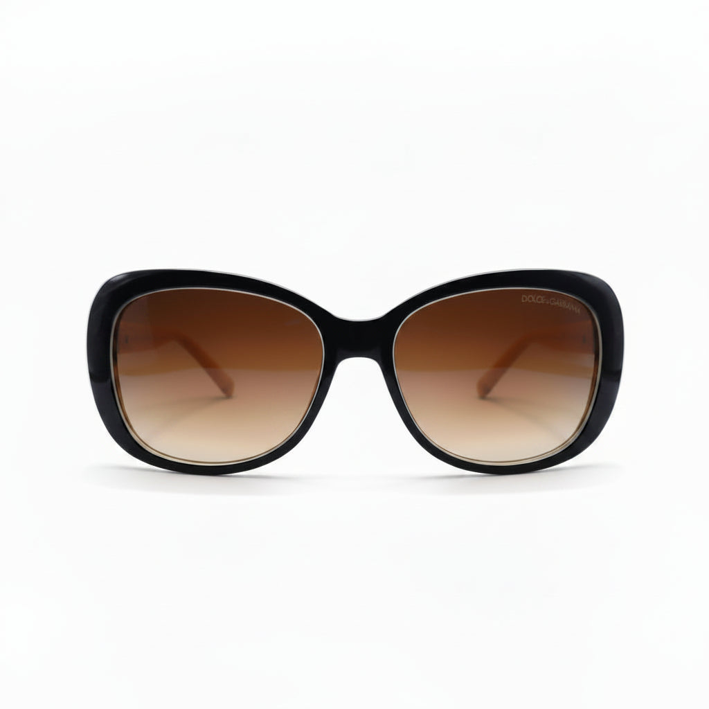 A pair of oversized cat-eye sunglasses with a glossy black frame and brown gradient lenses, displayed on a white surface with a reflective silver backdrop.