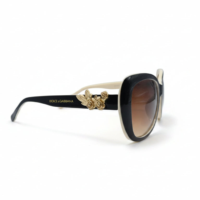 A pair of oversized cat-eye sunglasses with a glossy black frame and brown gradient lenses, displayed on a white surface with a reflective silver backdrop.