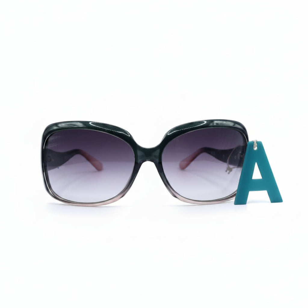 Oversized square sunglasses with a glossy black frame and gradient purple lenses, displayed on a white surface next to a blue letter 'A' prop, with textured black fabric bags on both sides.