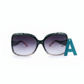 Oversized square sunglasses with a glossy black frame and gradient purple lenses, displayed on a white surface next to a blue letter 'A' prop, with textured black fabric bags on both sides.