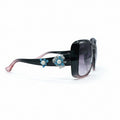 Black square sunglasses with pink-tipped arms, decorated with blue and white flower charms, shown on a white surface with black fabric props in the background.