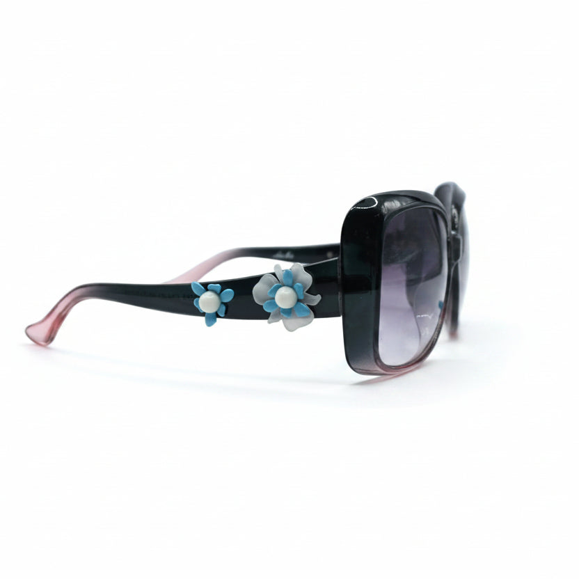 Black square sunglasses with pink-tipped arms, decorated with blue and white flower charms, shown on a white surface with black fabric props in the background.