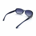 A pair of women's sunglasses with a dark gray acetate frame and gradient purple-blue lenses, displayed on a white surface with textured silver props in the background.