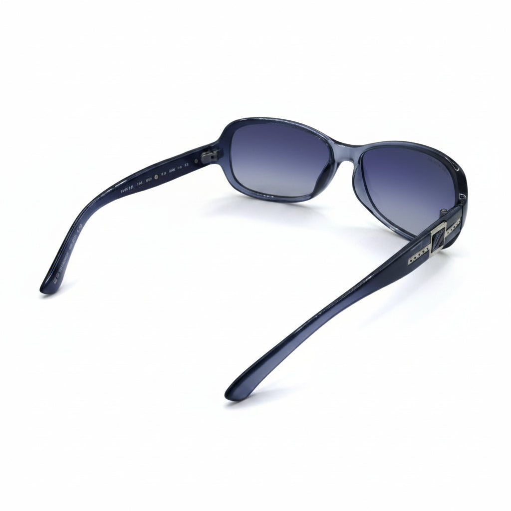 A pair of women's sunglasses with a dark gray acetate frame and gradient purple-blue lenses, displayed on a white surface with textured silver props in the background.