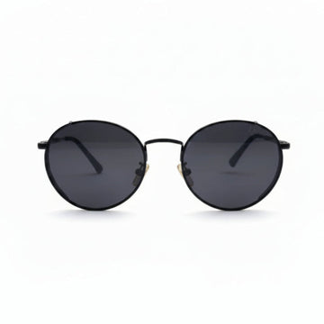 Round black sunglasses with dark lenses and a thin black metal frame, displayed on a white surface with two textured black bags on either side.