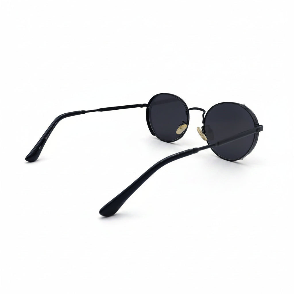 A pair of round black sunglasses with dark lenses and gold metal hinges, displayed on a white surface with two silver textured panels in the background.