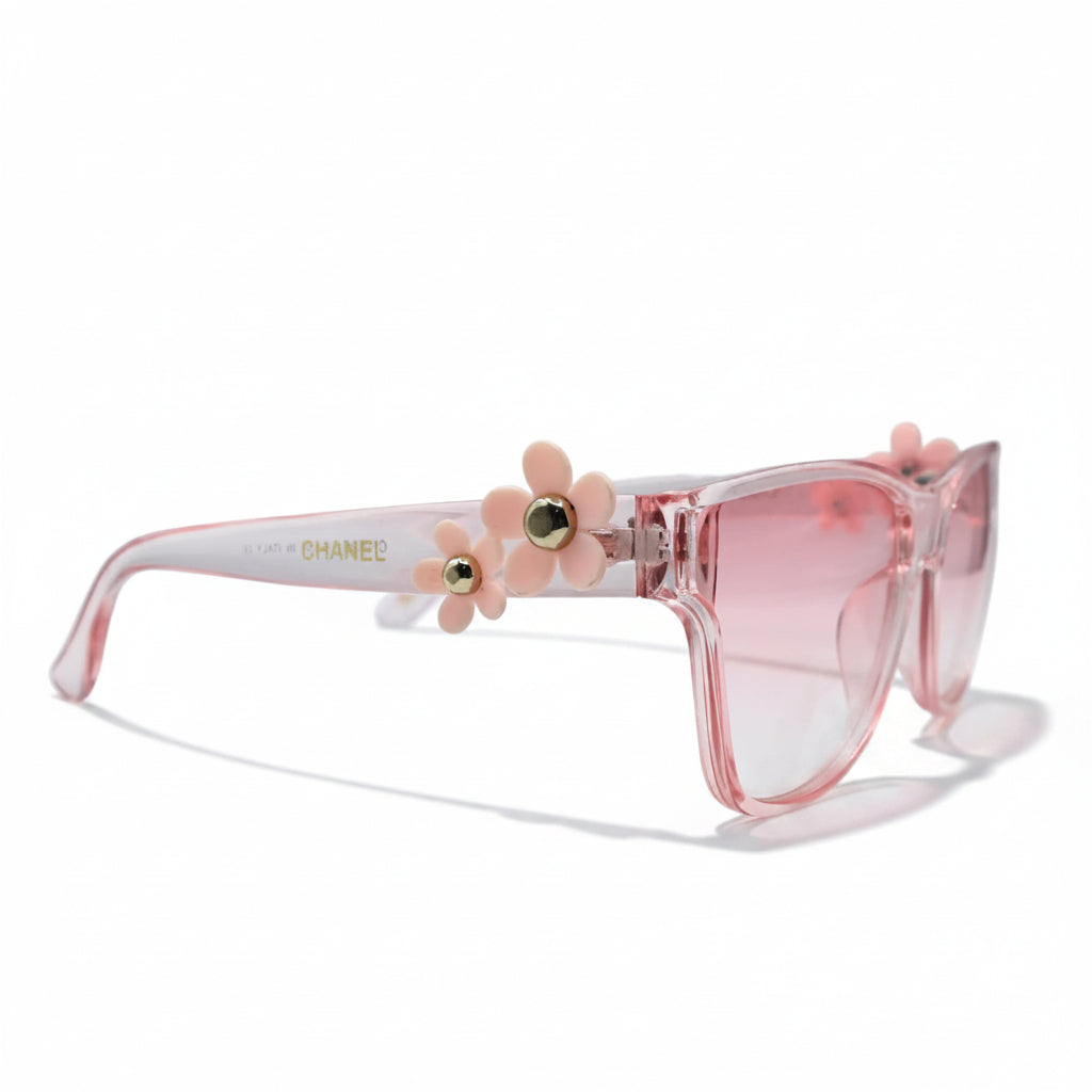 Pink cat-eye sunglasses with a translucent pink frame and pink gradient lenses, displayed on a white surface with black studio lights in the background.
