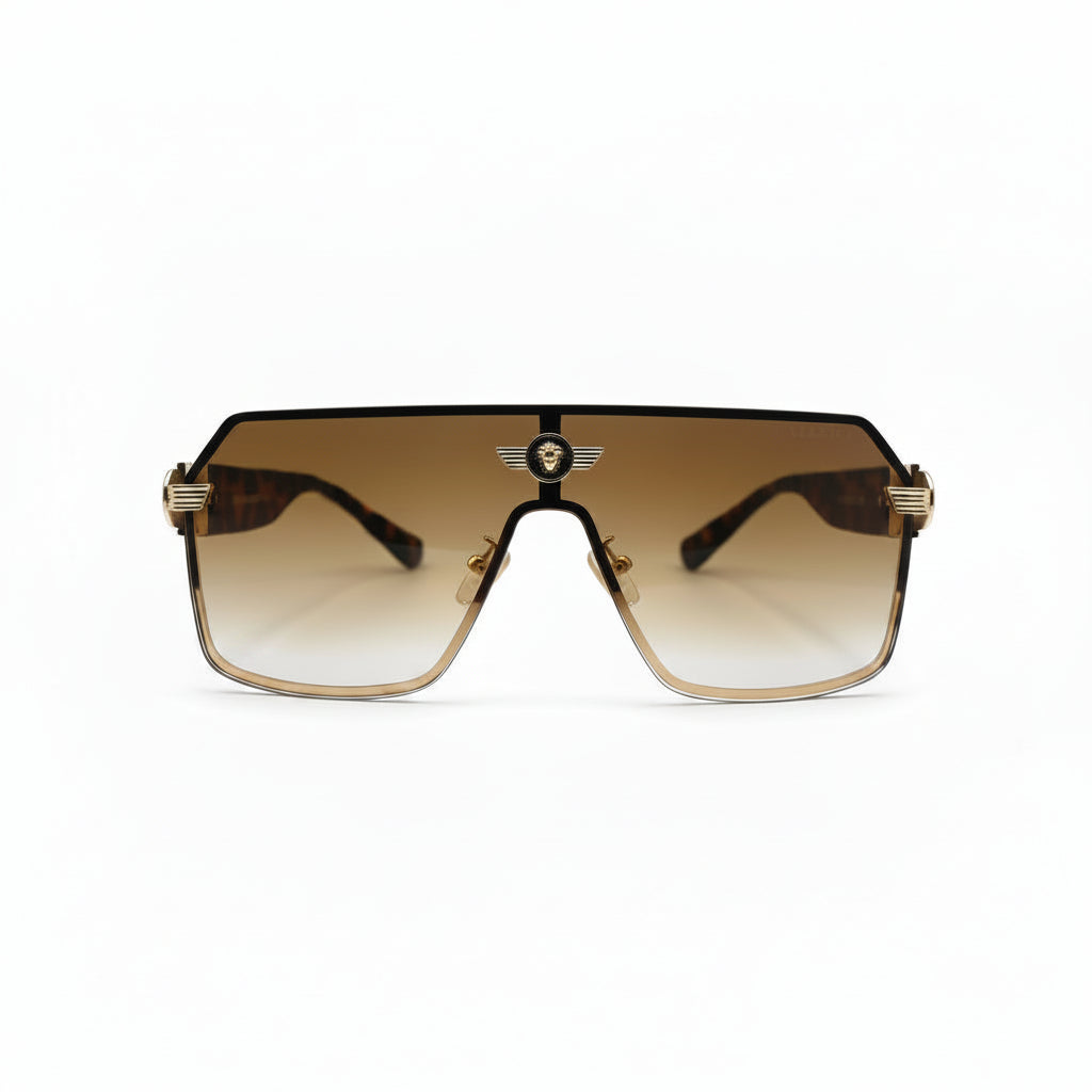 A pair of oversized shield-style sunglasses with a gradient brown lens and gold metal frames, displayed on a white surface with textured silver bags in the background.