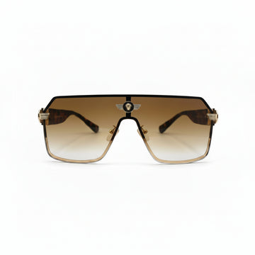 A pair of oversized shield-style sunglasses with a gradient brown lens and gold metal frames, displayed on a white surface with textured silver bags in the background.