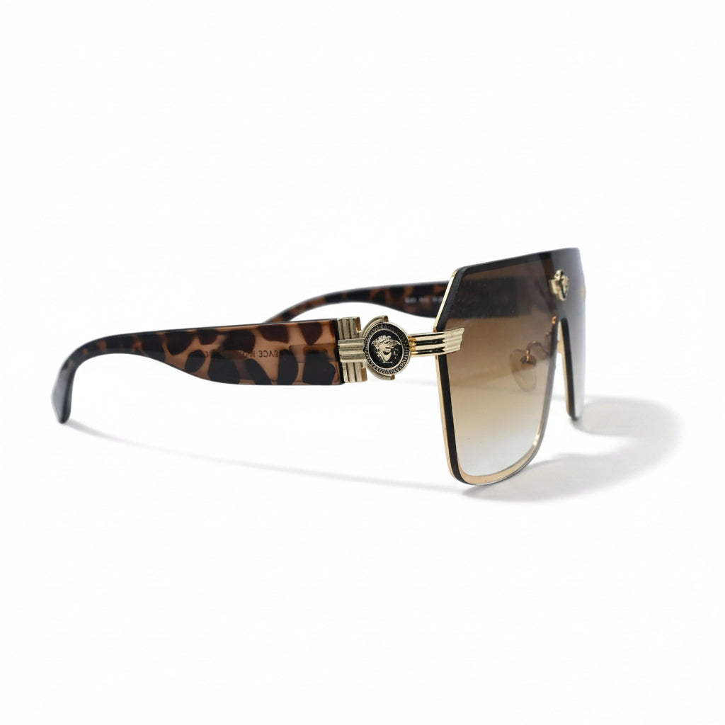 A pair of oversized shield-style sunglasses with a gradient brown lens and gold metal frames, displayed on a white surface with textured silver bags in the background.