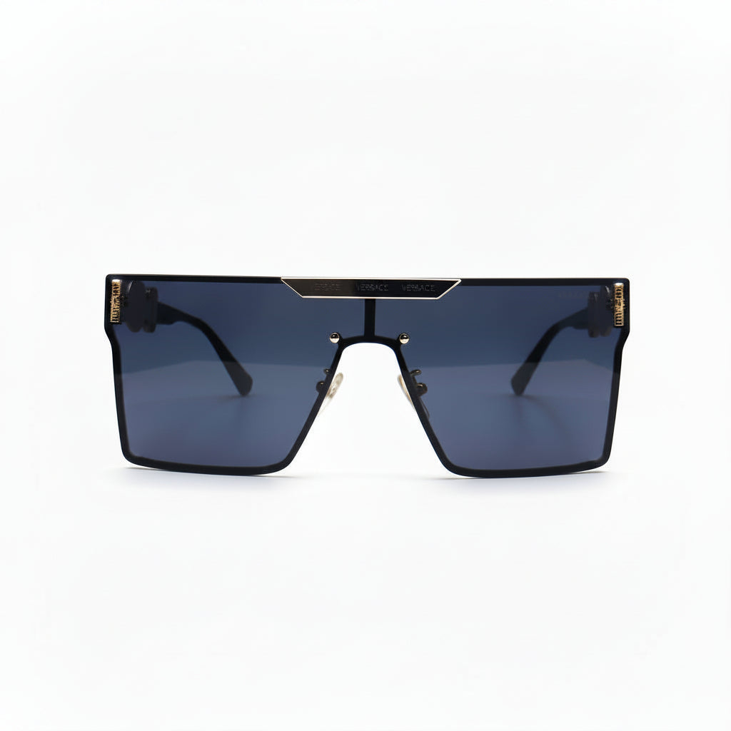 A pair of oversized shield-style sunglasses with a black frame, dark blue-gray lenses, and gold metal accents on the top bar and temples, displayed on a white surface with textured silver props on either side.