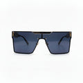 A pair of oversized shield-style sunglasses with a black frame, dark blue-gray lenses, and gold metal accents on the top bar and temples, displayed on a white surface with textured silver props on either side.