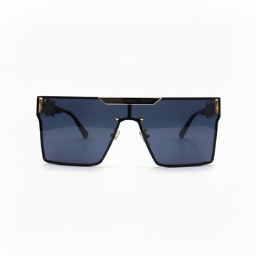 A pair of oversized shield-style sunglasses with a black frame, dark blue-gray lenses, and gold metal accents on the top bar and temples, displayed on a white surface with textured silver props on either side.