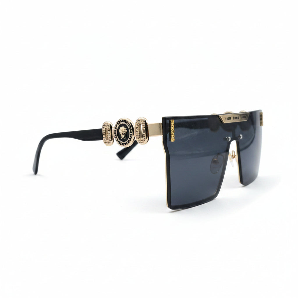 A pair of oversized shield-style sunglasses with a black frame, dark blue-gray lenses, and gold metal accents on the top bar and temples, displayed on a white surface with textured silver props on either side.