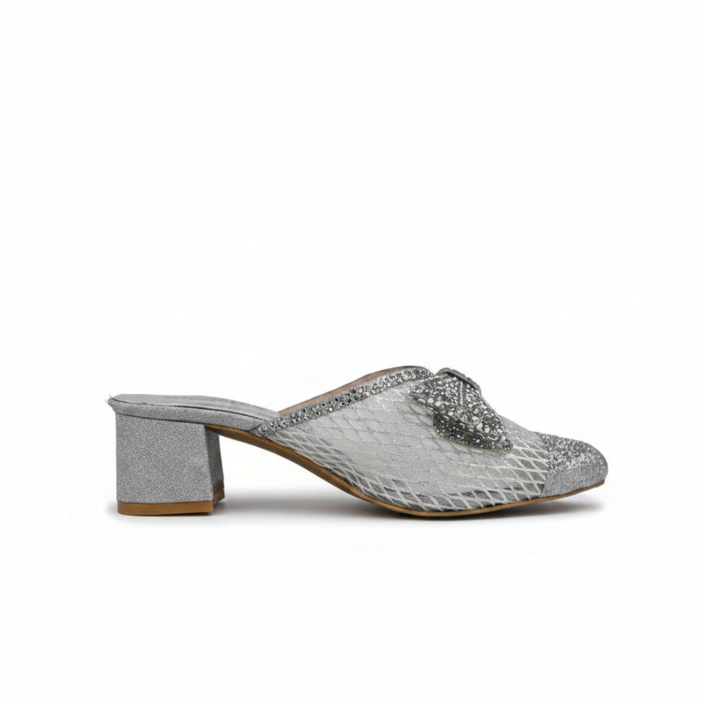 A single women's heeled mule sandal with a silver glittery snakeskin pattern, featuring a textured block heel and a decorative silver bow on the toe strap, set against a light gray background.