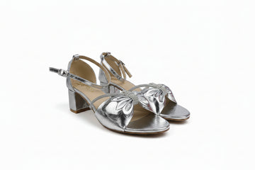 A pair of metallic silver strappy sandals with block heels, featuring a decorative bow embellishment on the toes and an ankle strap, shown on a white surface.