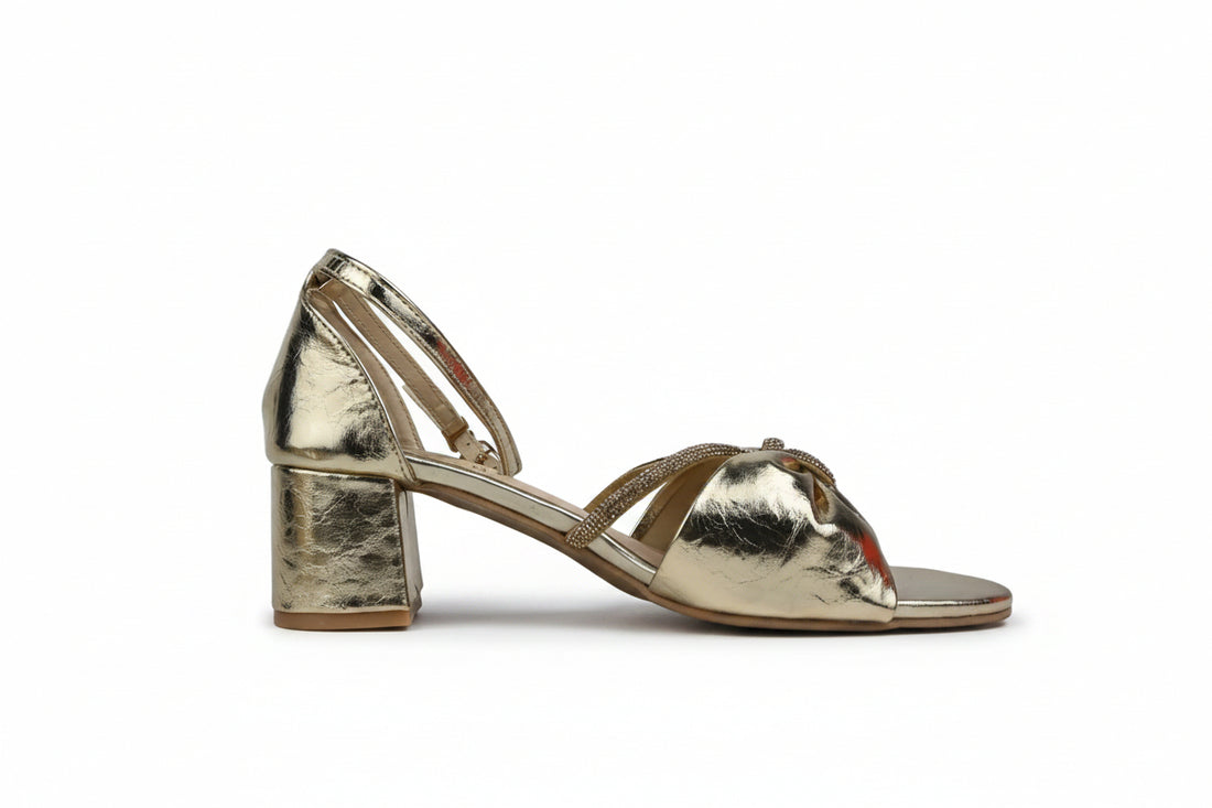 A pair of metallic silver strappy sandals with block heels, featuring a decorative bow embellishment on the toes and an ankle strap, shown on a white surface.