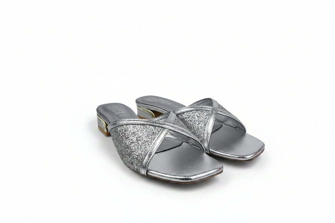 A pair of black leather slide sandals with textured glittery straps and low block heels with gold-toned detailing, photographed on a white fabric background.