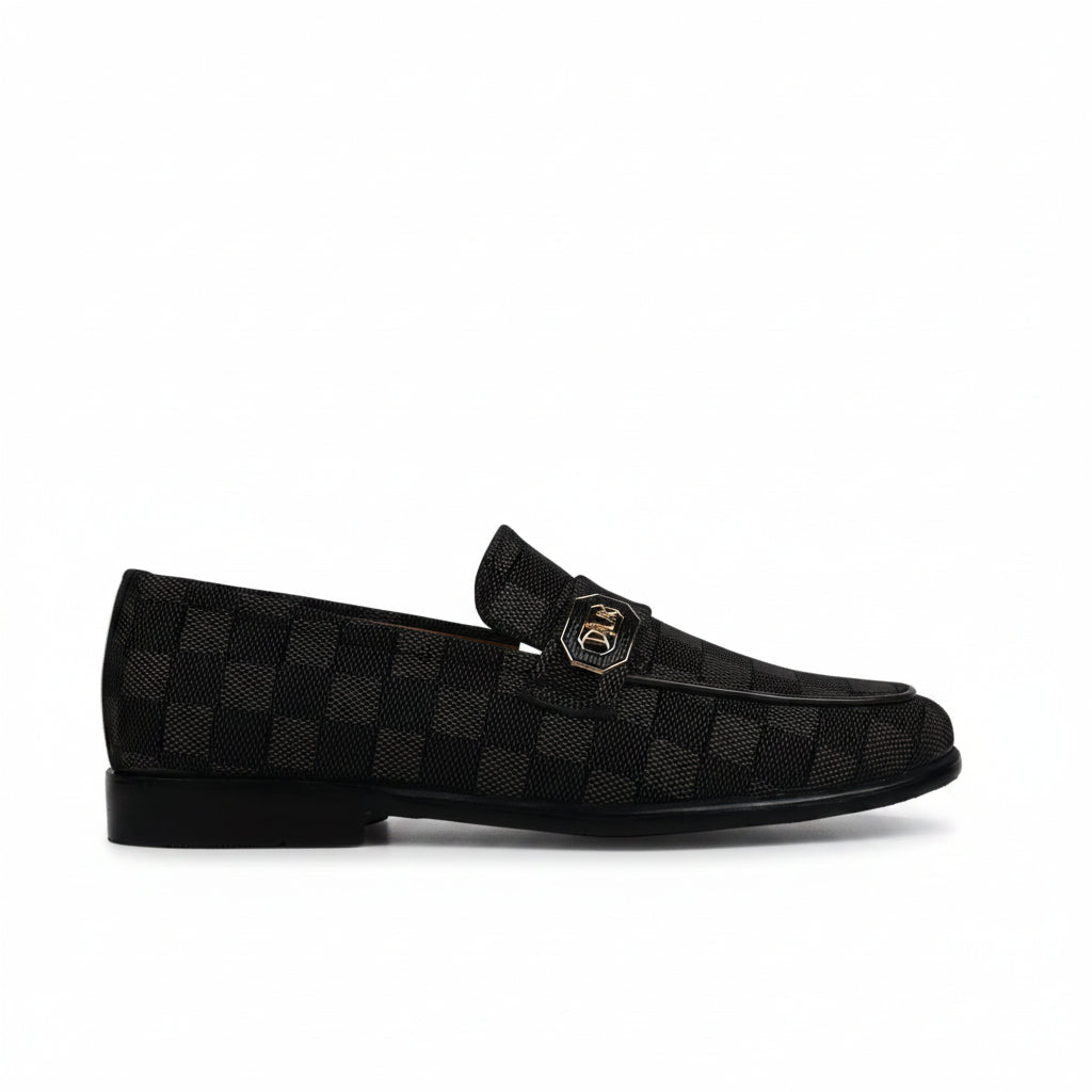 A single black checkered loafer with a small gold-tone metal emblem on the vamp, photographed against a wrinkled white studio backdrop.