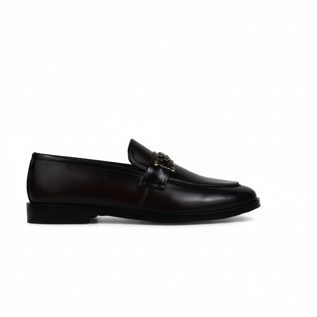 A single dark brown leather penny loafer with a decorative strap and small gold-tone buckle detail, positioned horizontally against a wrinkled white studio backdrop.